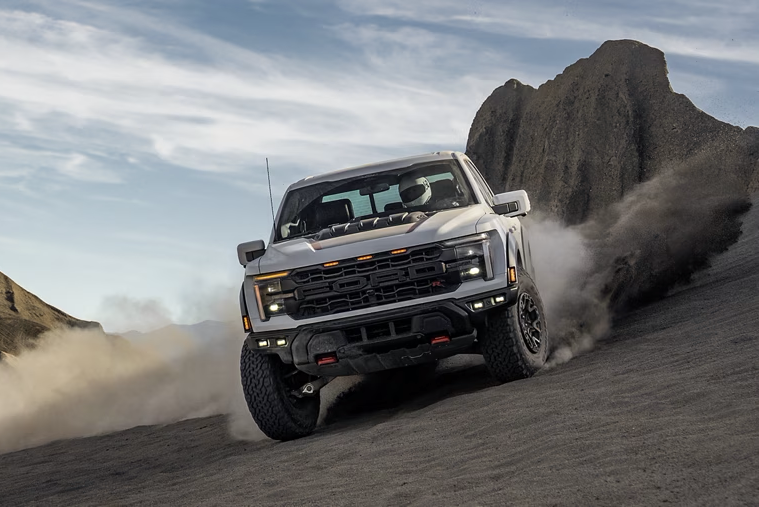 A white Ford Raptor pickup truck is driving uphill on a sandy or rocky off-road terrain, kicking up dirt and dust, with a rugged landscape in the background under a cloudy sky.
