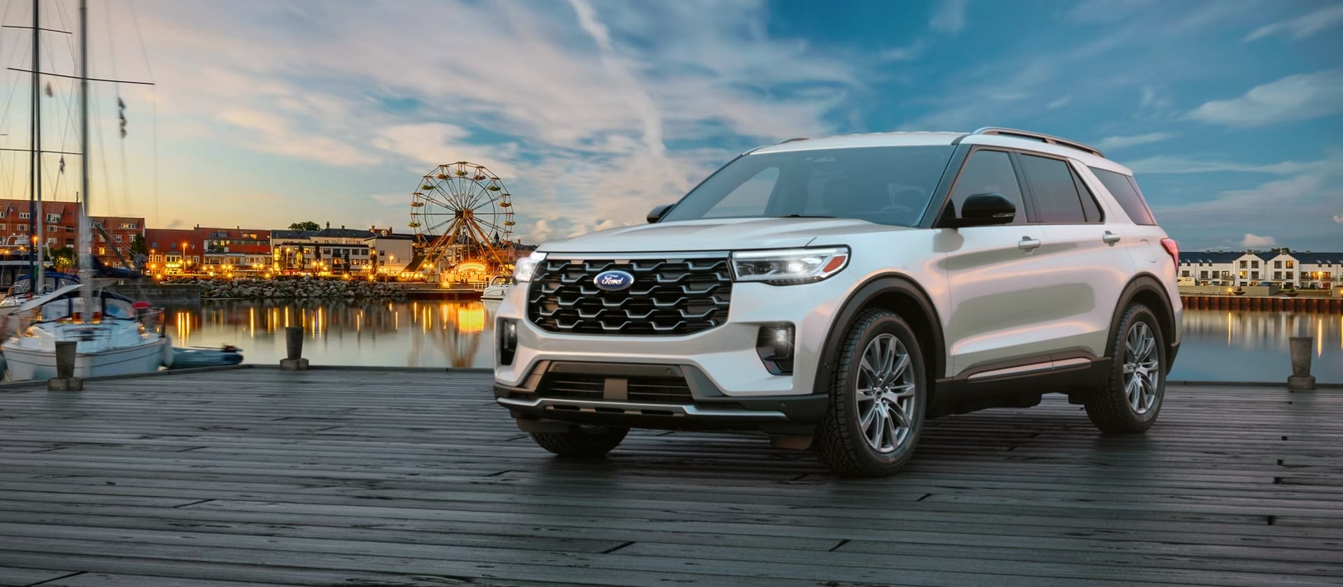 A silver 2026 Ford Explorer in Chandler AZ is parked facing the viewer to the left on a dock by a calm bay at dusk, with a lively seaside village in the background.