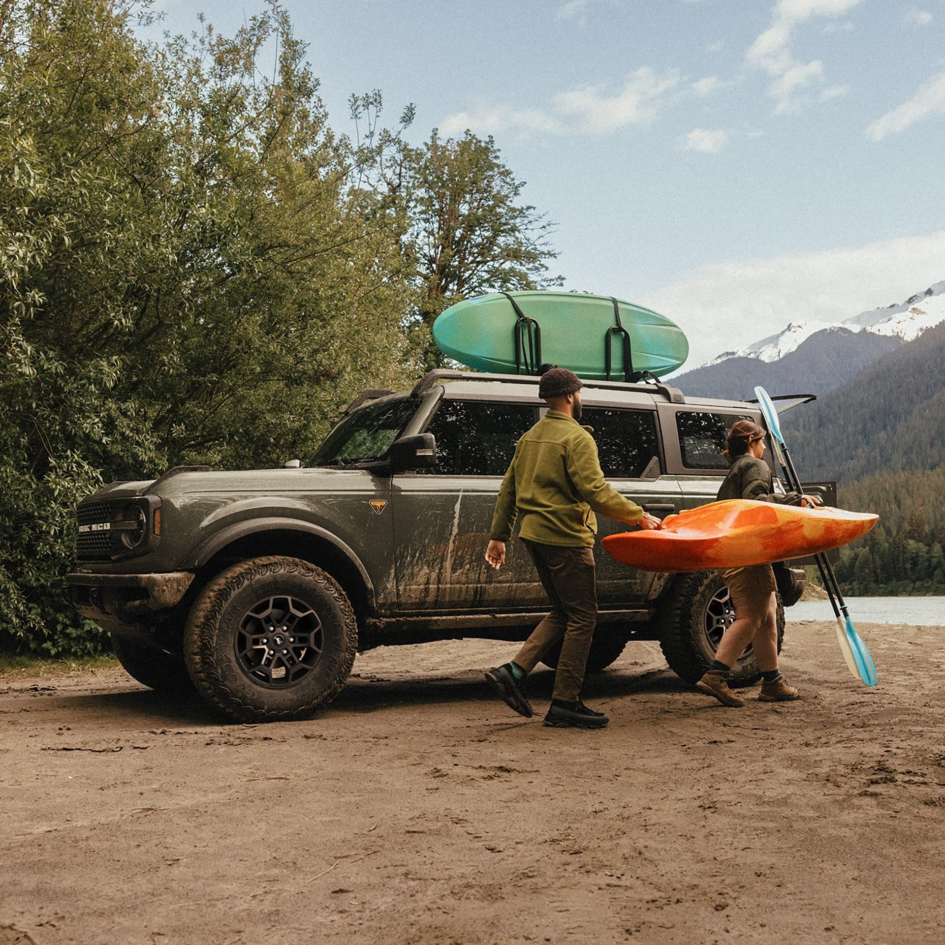 A grey 2026 Ford Bronco Sport in Phoenix is parked facing left on a riverbank with tree-covered mountains in the background. A green kayak is attached to the top of the Ford offroad SUV. Two people are carrying an orange kayak and a blue paddle to the right towards the river. The 2026 Bronco Sport is covered in mud.